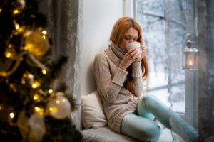 Young beautiful woman with reddish hair sitting home by the window with cup of hot coffee wearing knitted warm sweater. Christmas tree with decorations and lights in the room, snowy winter outside